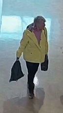 woman with short grey hair wearing a bright yellow jacket and black pants. She is carrying two black bags, one in each hand. 