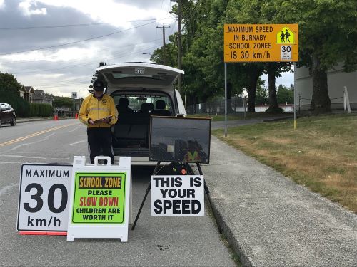 A volunteer in a yellow jacket stands beside a vehicle with its trunk open and several signs related to slowing down in school zones