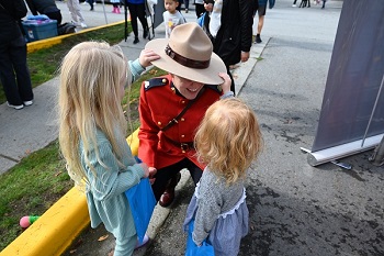 A Mountie in red serge kneeing down while two little girl’s play with the mountie’s Stetson 