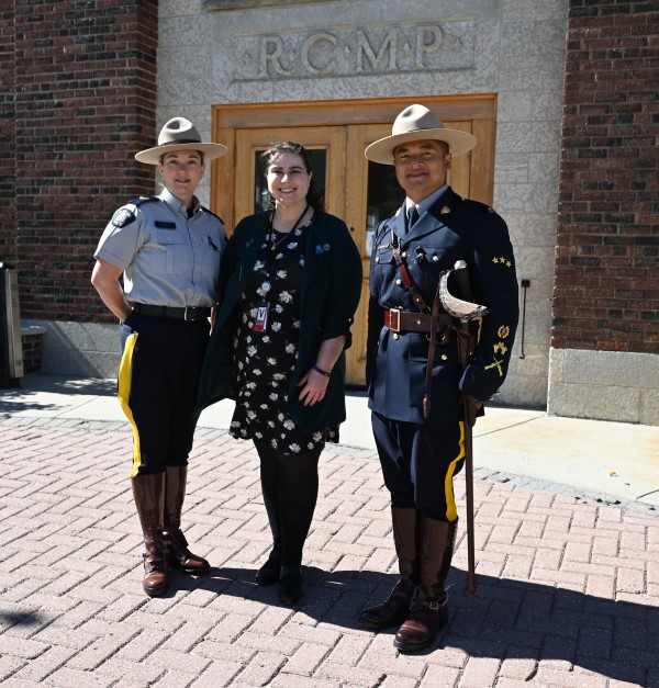 Burnaby RCMP Cpl. Laura Hirst poses with Burnaby RCMP Municipal Employee Katina Stachura and Staff Sgt. Major David Douangchanh in front of the brick drill hall at Depot Division in Regina. All three are smiling, 