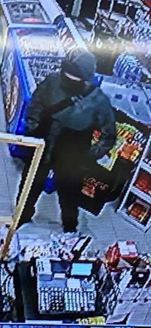 The male suspect wearing a black and green jacket with the hood up, black pants, a mask covering his face, and a duffle bag strap across his chest.