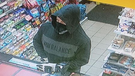 Un homme de race blanche qui porte un masque noir et un chandail noir arborant le nom de marque « New Balance » se tient devant un comptoir dans un dépanneur. 