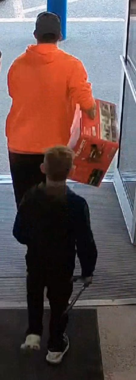 Image de dos du suspect vêtu d’un chandail orange qui transporte une boîte à l’extérieur du commerce. Il est suivi d’un enfant portant des vêtements foncés.