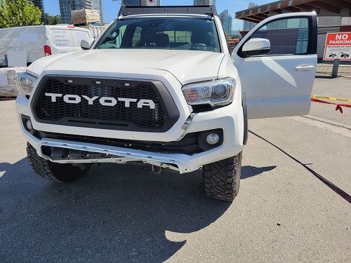 Une Toyota Tacoma blanche dont l’avant est endommagé et dont la portière du conducteur est ouverte. Le mot TOYOTA est écrit en blanc sur la calandre noire de la camionnette.