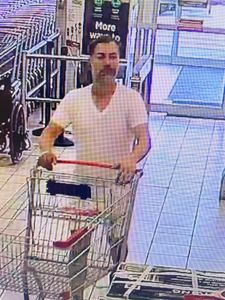 third male suspect pushing a cart into the store. He is wearing a white v-neck tshirt and light coloured shorts. He has short dark hair and a greying beard or 5 o’clock shadow. 