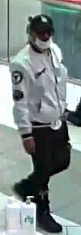 A man walking to the right wearing a dark LA Dodgers baseball hat, white medical mask, white bomber jacket, black pants and dark shoes.