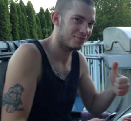 Kyle in a black tank top, exposing an animal tattoo on his right upper arm. His hair is buzzed. 