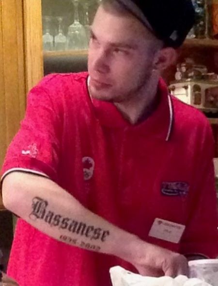 Kyle in a black hat and red short-sleeved shirt, showing the ‘Bassanese’ tattoo on his right forearm. 